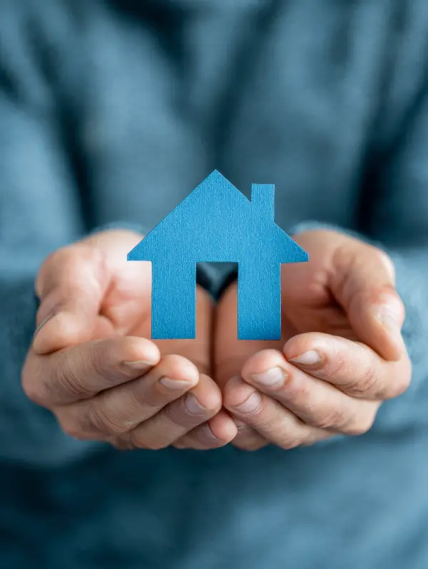 Une main tient un petit parapluie bleu protégeant une maison miniature pour symboliser l'assurance