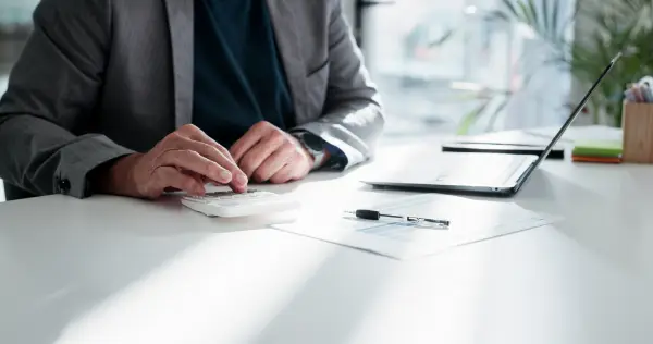 Plan serré sur des mains posées sur un bureau avec un ordinateur, une calculatrice et un dossier