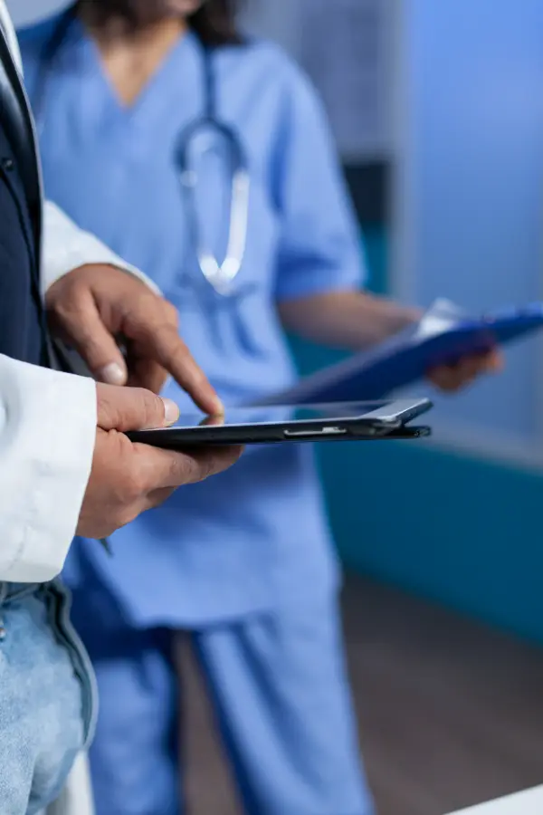 Plan très serré sur les mains de médecin avec une tablette. En arrière plan, un autre médecin en blouse bleue avec une dossier